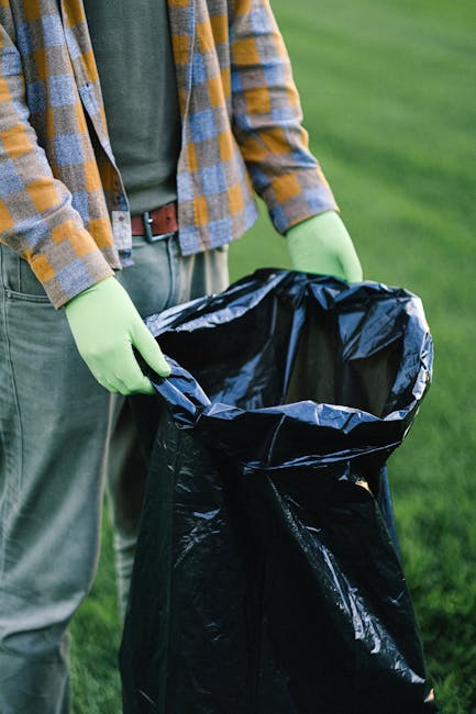 In the foreground, a large pile of mixed waste materials includes cardboard boxes, plastic containers, and discarded paper, all jumbled together on a paved driveway. The cardboard boxes vary in size, 